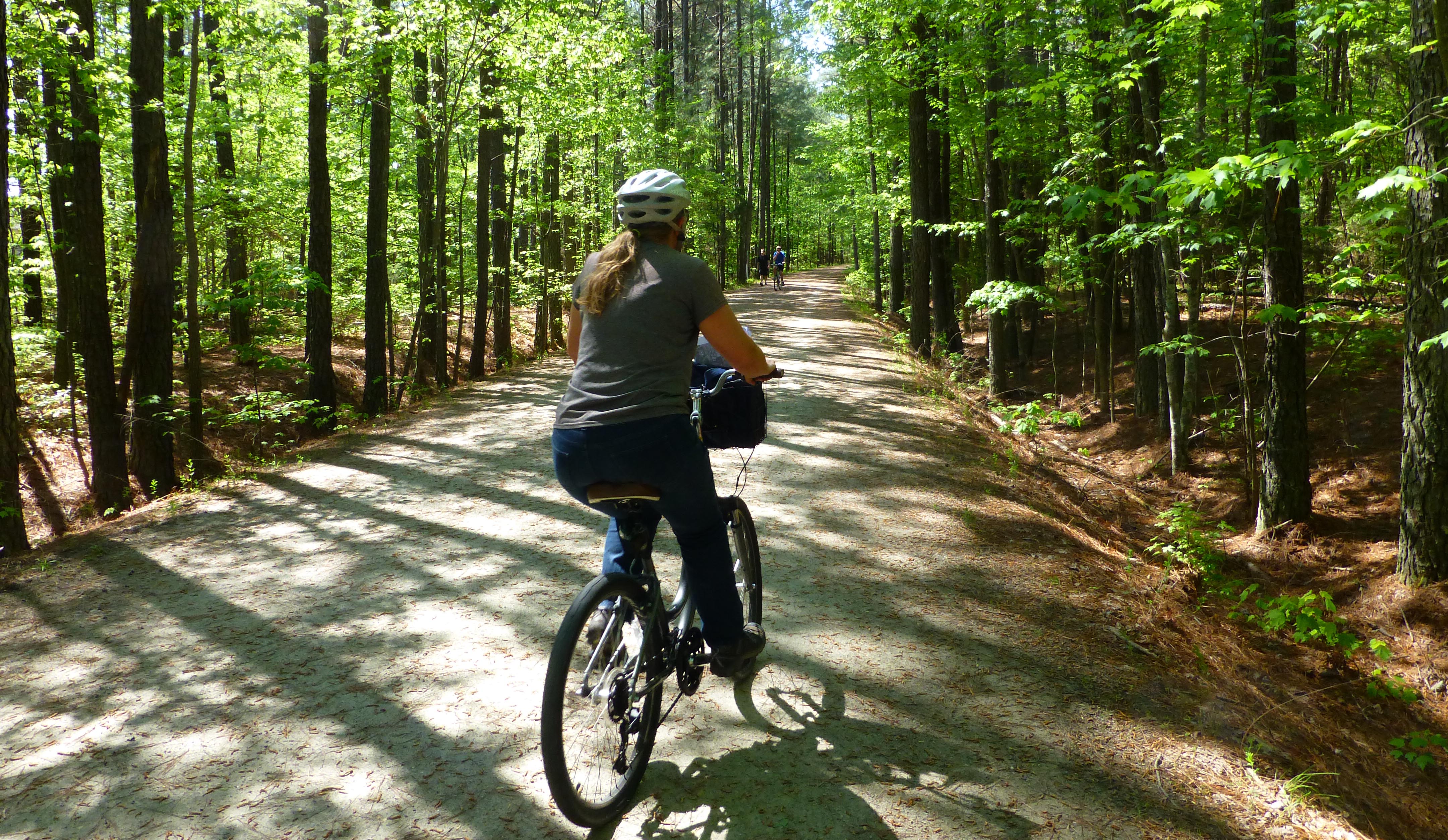 A woman rides her bike on a path surrounded by woods. 