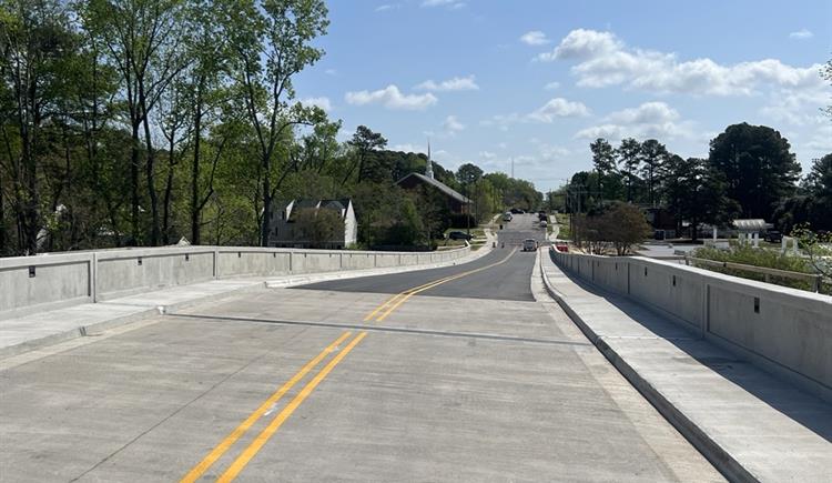There is a bridge with yellow solid lines with green trees in the back ground and a bright blue sky.