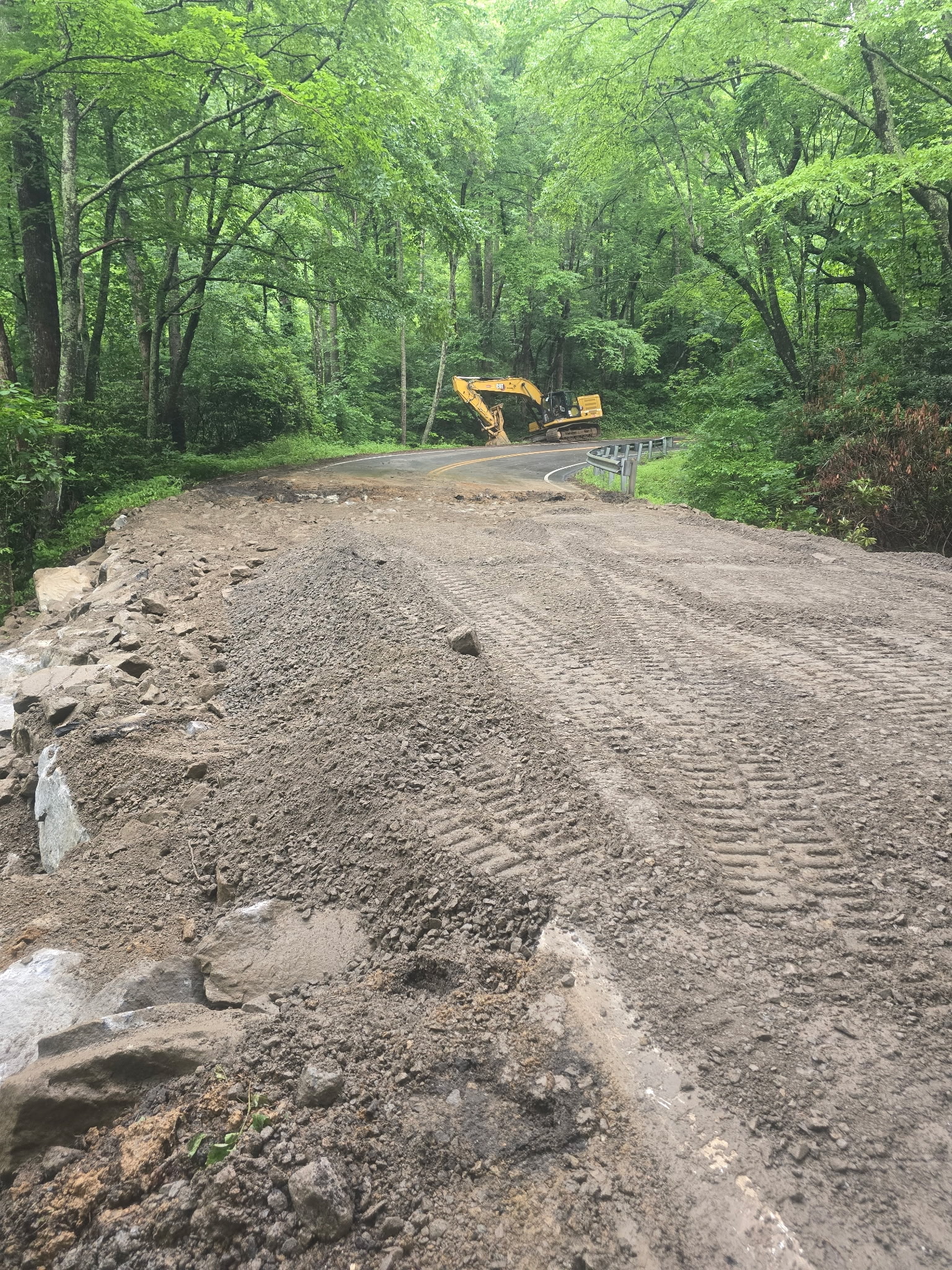 An excavator sits off the road behind a worksite in a wooded area. 